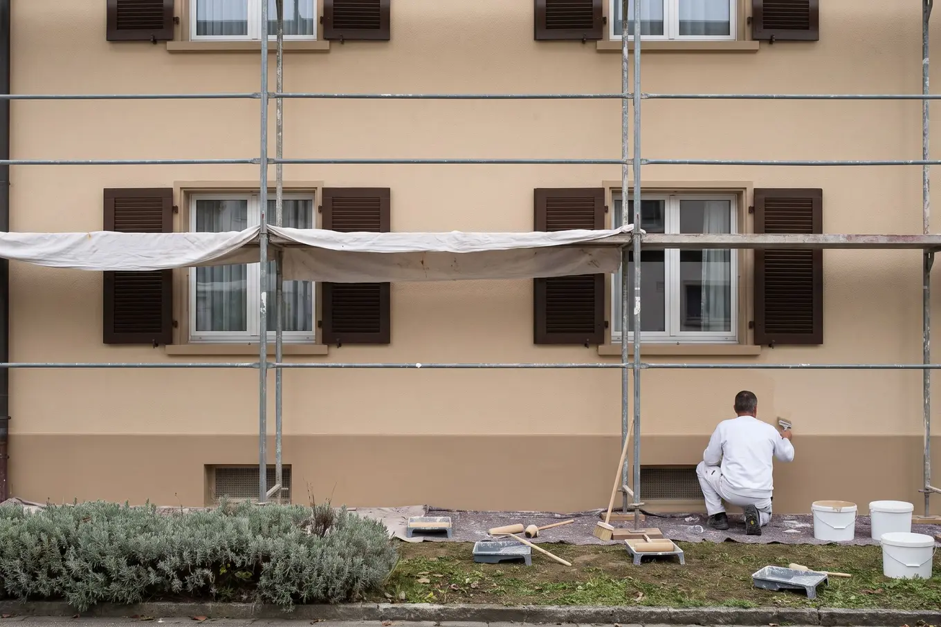 Frisch gestrichene Fassade mit Gerüst und dunklen Fensterläden, ein Maler von hinten arbeitet am Sockel